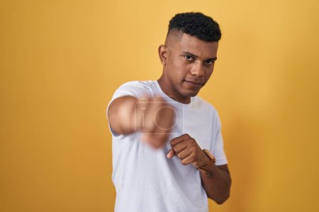 Foto de Joven hispano de pie sobre fondo amarillo golpeando puño para luchar, ataque agresivo y enojado, amenaza y violencia - Imagen libre de derechos