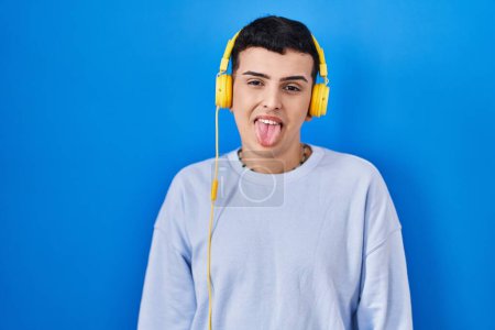 Foto de Persona no binaria escuchando música usando auriculares que sacan la lengua feliz con expresión divertida. concepto de emoción. - Imagen libre de derechos
