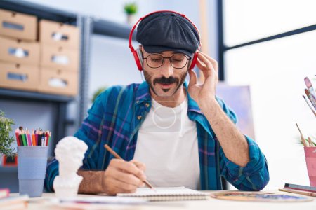 Photo for Young hispanic man artist listening to music drawing on notebook at art studio - Royalty Free Image