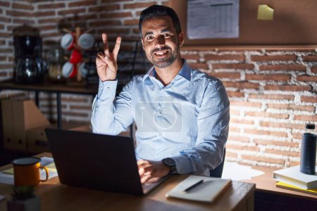 Téléchargez les photos : Homme hispanique avec la barbe travaillant au bureau la nuit souriant en regardant la caméra montrant des doigts faisant signe de victoire. numéro deux. - en image libre de droit