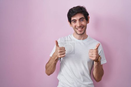 Foto de Young hispanic man standing over pink background success sign doing positive gesture with hand, thumbs up smiling and happy. cheerful expression and winner gesture. - Imagen libre de derechos
