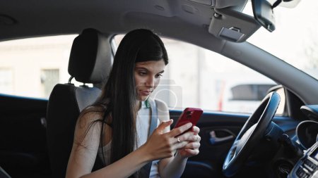 Foto de Young beautiful hispanic woman using smartphone sitting on car at street - Imagen libre de derechos