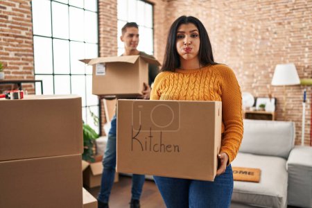 Photo for Young couple moving to a new home with cardboard boxes puffing cheeks with funny face. mouth inflated with air, catching air. - Royalty Free Image