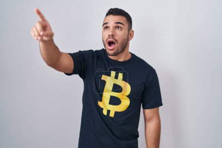 Photo for Young hispanic man wearing bitcoin t shirt pointing with finger surprised ahead, open mouth amazed expression, something on the front - Royalty Free Image