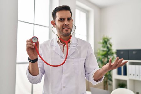 Photo for Young hispanic doctor man with beard holding stethoscope auscultating clueless and confused expression. doubt concept. - Royalty Free Image