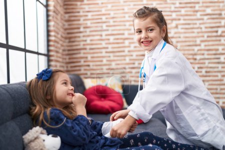 Photo for Adorable girls wearing doctor uniform bandaging arm at home - Royalty Free Image