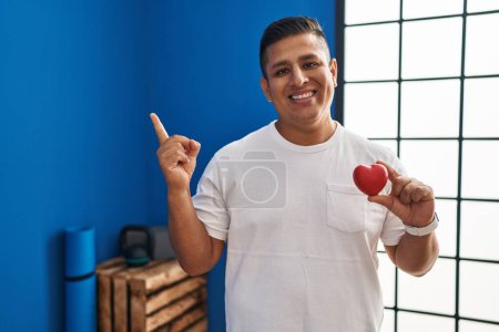 Téléchargez les photos : Jeune homme hispanique tenant coeur rouge à la salle de gym souriant heureux pointant avec la main et le doigt sur le côté - en image libre de droit