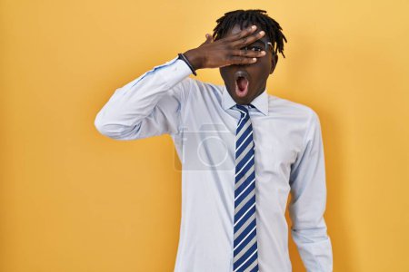 Foto de Hombre africano con rastas de pie sobre fondo amarillo asomándose en shock cubriendo la cara y los ojos con la mano, mirando a través de los dedos con expresión avergonzada. - Imagen libre de derechos