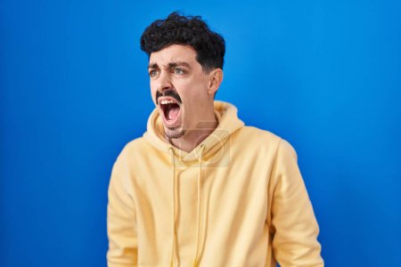 Photo for Hispanic man standing over blue background angry and mad screaming frustrated and furious, shouting with anger. rage and aggressive concept. - Royalty Free Image