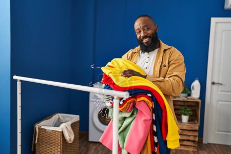 Foto de Joven afroamericano hombre sonriendo confiado sosteniendo ropa en la sala de lavandería - Imagen libre de derechos