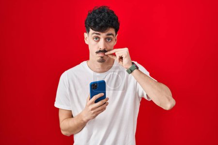 Téléchargez les photos : Homme hispanique utilisant smartphone sur fond rouge bouche et lèvres fermées comme zip avec les doigts. secret et silencieux, tabou parlant - en image libre de droit
