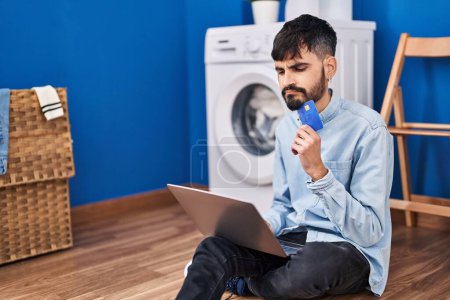 Téléchargez les photos : Young hispanic man using laptop and credit card waiting for washing machine at laundry room - en image libre de droit