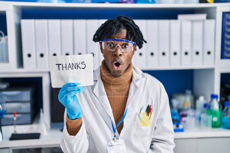 Foto de Joven africano con rastas trabajando en laboratorio científico asustado y sorprendido con la boca abierta para sorpresa, cara de incredulidad - Imagen libre de derechos