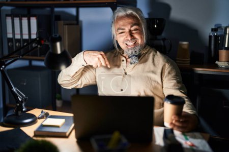 Foto de Hombre de mediana edad con el pelo gris trabajando en la oficina por la noche mirando confiado con sonrisa en la cara, señalándose con los dedos orgullosos y felices. - Imagen libre de derechos