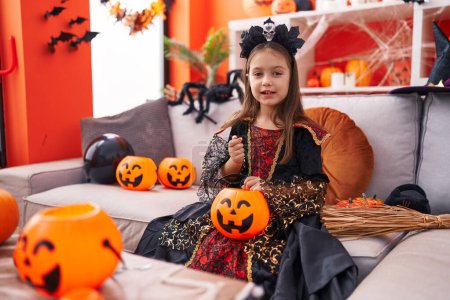 Foto de Adorable chica hispana con disfraz de halloween sosteniendo dulces de canasta de calabaza en casa - Imagen libre de derechos