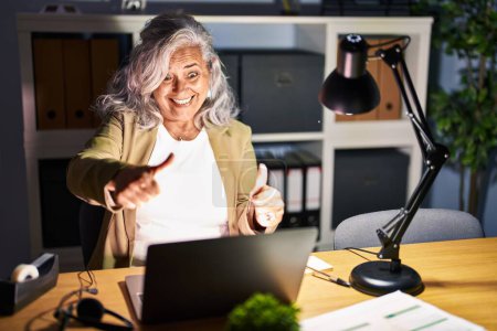 Foto de Mujer de mediana edad con el pelo gris trabajando con computadora portátil tarde en la noche aprobando hacer gesto positivo con la mano, pulgares arriba sonriendo y feliz por el éxito. gesto ganador. - Imagen libre de derechos