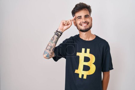 Foto de Joven hombre hispano con tatuajes con camiseta bitcoin sonriendo apuntando a la cabeza con un dedo, gran idea o pensamiento, buena memoria - Imagen libre de derechos