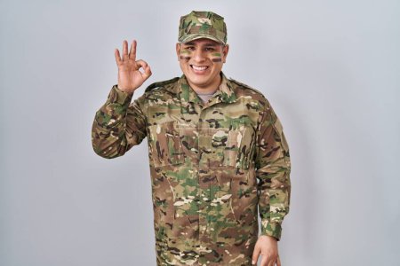 Téléchargez les photos : Jeune homme hispanique portant uniforme de camouflage armée souriant positif faire ok signe avec la main et les doigts. expression réussie. - en image libre de droit