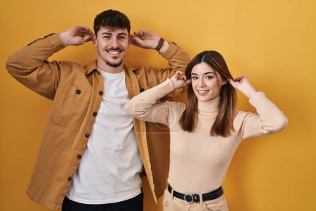 Foto de Joven pareja hispana de pie sobre fondo amarillo sonriendo tirando de las orejas con los dedos, gesto divertido. problema de audición - Imagen libre de derechos