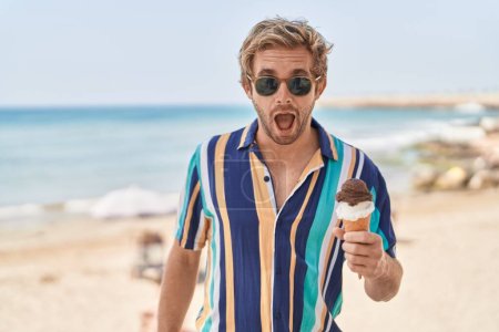 Foto de Hombre caucásico comiendo un helado en la playa asustado y sorprendido con la boca abierta para sorpresa, cara de incredulidad - Imagen libre de derechos
