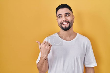 Photo for Young handsome man wearing casual t shirt over yellow background smiling with happy face looking and pointing to the side with thumb up. - Royalty Free Image