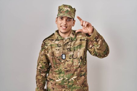 Téléchargez les photos : Jeune homme arabe portant uniforme camouflage armée geste souriant et confiant avec la main faisant signe de petite taille avec les doigts à la recherche et la caméra. concept de mesure. - en image libre de droit