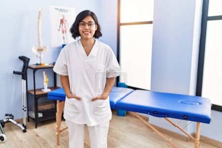 Téléchargez les photos : Jeune femme latine portant un uniforme de physiothérapeute debout à la clinique de physiothérapie - en image libre de droit