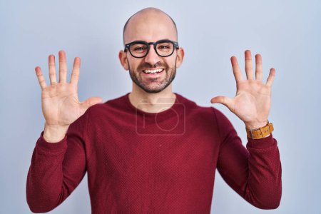 Foto de Joven hombre calvo con barba de pie sobre fondo blanco con gafas que muestran y señalan con los dedos número diez mientras sonríe confiado y feliz. - Imagen libre de derechos