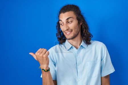 Photo for Young hispanic man standing over blue background smiling with happy face looking and pointing to the side with thumb up. - Royalty Free Image