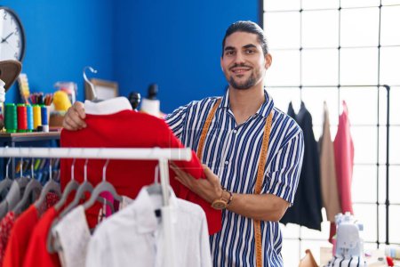 Téléchargez les photos : Young hispanic man tailor smiling confident holding t shirt on rack at sewing studio - en image libre de droit