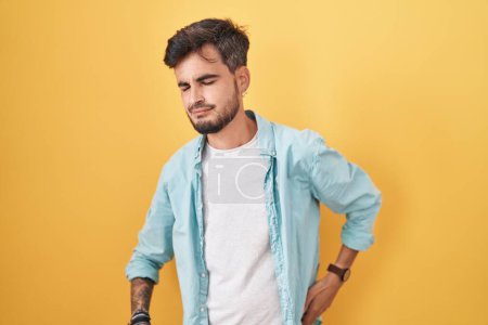 Téléchargez les photos : Jeune homme hispanique tatoué debout sur fond jaune souffrant de maux de dos, touchant le dos avec la main, douleurs musculaires - en image libre de droit