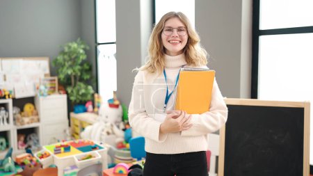 Téléchargez les photos : Young blonde woman preschool teacher smiling confident holding books at kindergarten - en image libre de droit