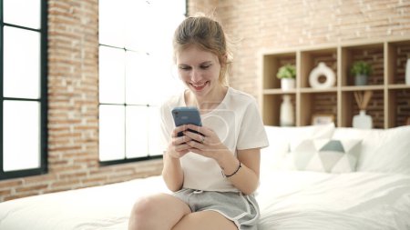 Téléchargez les photos : Jeune femme blonde utilisant smartphone assis sur le lit dans la chambre - en image libre de droit