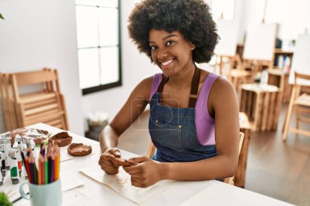 Téléchargez les photos : Jeune femme afro-américaine souriante confiante manipulant de l'argile au studio d'art - en image libre de droit