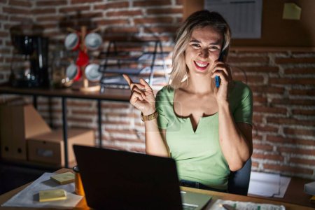 Photo for Young beautiful woman working at the office at night speaking on the phone with a big smile on face, pointing with hand finger to the side looking at the camera. - Royalty Free Image