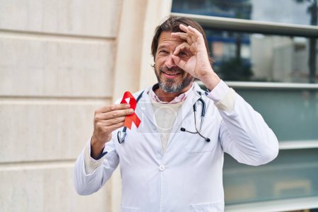 Photo for Handsome middle age doctor man holding support red ribbon smiling happy doing ok sign with hand on eye looking through fingers - Royalty Free Image