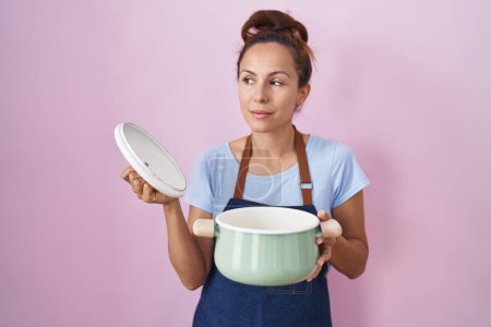 Téléchargez les photos : Brunette femme portant tablier tenant casserole souriant regardant vers le côté et regardant loin en pensant. - en image libre de droit