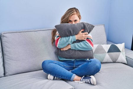 Téléchargez les photos : Young blonde woman hugging cushion sitting on sofa at home - en image libre de droit