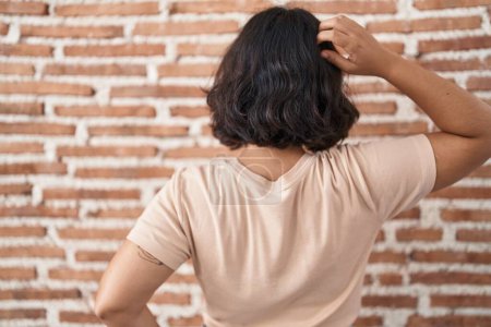 Téléchargez les photos : Jeune femme hispanique debout sur le mur de briques vers l'arrière en pensant au doute avec la main sur la tête - en image libre de droit