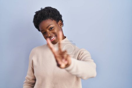 Téléchargez les photos : Femme afro-américaine debout sur fond bleu souriant en regardant la caméra montrant les doigts faisant signe de victoire. numéro deux. - en image libre de droit