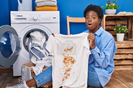 Téléchargez les photos : Femme afro-américaine tenant t-shirt avec tache sale peur et choqué avec surprise et expression étonnante, peur et visage excité. - en image libre de droit