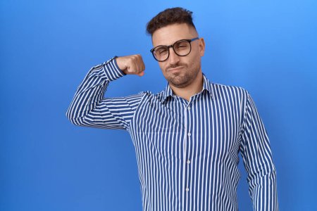 Téléchargez les photos : Homme hispanique avec barbe portant des lunettes personne forte montrant muscle du bras, confiant et fier de la puissance - en image libre de droit