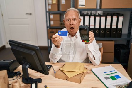 Photo for Senior man with grey hair working at small business ecommerce holding credit card and dataphone afraid and shocked with surprise and amazed expression, fear and excited face. - Royalty Free Image
