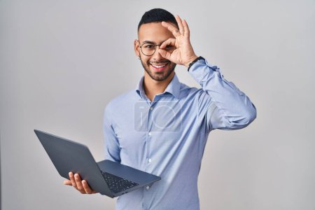 Foto de Joven hombre hispano trabajando usando computadora portátil haciendo buen gesto con la mano sonriendo, los ojos mirando a través de los dedos con la cara feliz. - Imagen libre de derechos