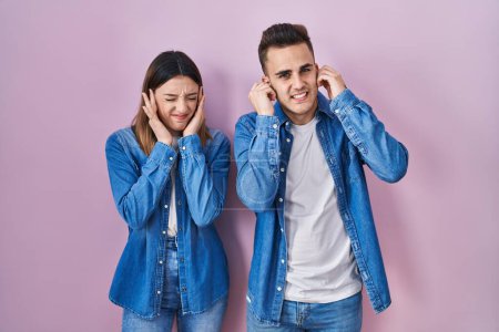 Téléchargez les photos : Jeune couple hispanique debout sur fond rose couvrant les oreilles avec des doigts avec une expression agacée pour le bruit de la musique forte. concept sourd. - en image libre de droit