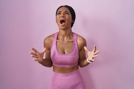 Foto de Mujer afroamericana con trenzas que usan ropa deportiva sobre fondo rosa loca y loca gritando y gritando con expresión agresiva y los brazos levantados. concepto de frustración. - Imagen libre de derechos