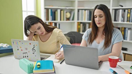Téléchargez les photos : Deux femmes assises sur la table étudiant en utilisant des notes d'écriture d'ordinateur portable à l'université de bibliothèque - en image libre de droit