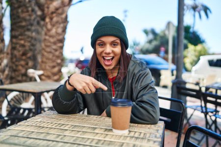 Foto de Mujer hispana joven bebiendo una taza de café al aire libre sonriendo feliz señalando con la mano y el dedo - Imagen libre de derechos