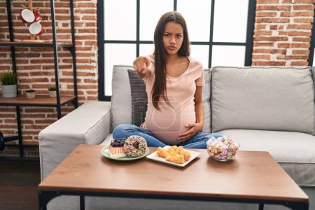 Photo for Young pregnant woman eating sweets at home pointing with finger to the camera and to you, confident gesture looking serious - Royalty Free Image