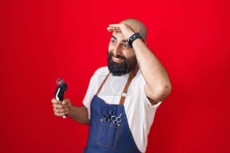 Photo for Young hispanic man with beard and tattoos wearing barber apron holding razor very happy and smiling looking far away with hand over head. searching concept. - Royalty Free Image